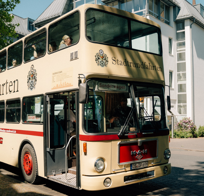 Doppeldeckerbus in Cremefarben, mit Passagieren am Einstieg, vor modernen Gebäuden.