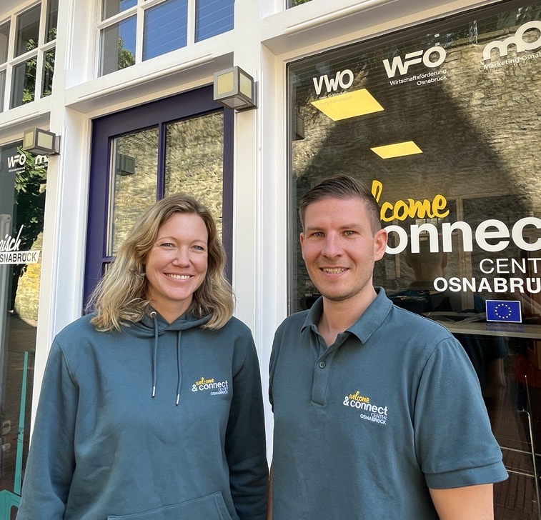 Zwei Personen sitzen auf Stühlen vor einem Gebäude mit bunten Fenstern und einem Schild.